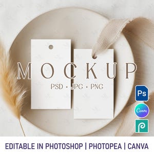 Könnte beinhalten: Zwei weiße rechteckige Anhänger mit einem Loch oben, beschriftet mit "MOCKUP" und darunter "PSD + JPG + PNG". Die Anhänger liegen auf einem beigen Teller mit einem Band und einer getrockneten Pflanze. Der Text "EDITABLE IN PHOTOSHOP | PHOTOPEA | CANVA" steht unten.