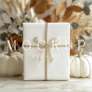 May include: A white gift box with a beige satin ribbon tied in a bow. The box is sitting on a table with two white pumpkins in the background.