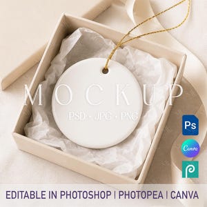 May include: A round white ceramic ornament with a hole at the top for hanging, featuring a gold string. The ornament is nestled inside a square beige gift box lined with white tissue paper. Text on the image reads MOCKUP PSD + JPG + PNG.
