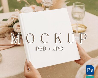 May include: A white book with the word "MOCKUP" and "PSD + JPG" printed on the cover. The book is held by a person. The background includes a table, flowers, and a wine glass.