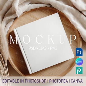 May include: A white square book mockup with the word "MOCKUP" in large white letters. The book is on a wooden tray with a beige cloth and decorative grass. Text on the book says "PSD + JPG + PNG". Icons for Photoshop, Canva, and Photopea are also visible.