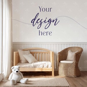 May include: A light wood crib with bedding and pillows, a wicker chair with a cushion, and a plush white teddy bear on a rug. The wall has the text "Your design here" in navy blue script.