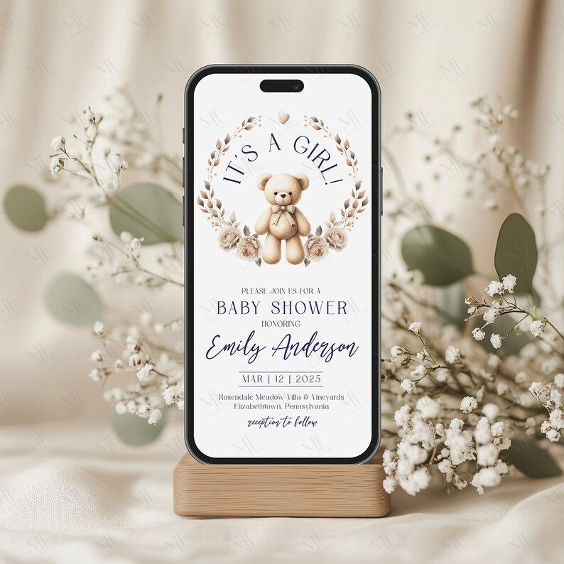 Op de afbeelding: Een digitale uitnodiging voor een babyshower met een illustratie van een teddybeer en een bloemenkrans. De tekst luidt "It's a Girl!" en bevat de naam "Emily Anderson", de datum "Mar 12 | 2025" en de locatie "Rosendale Meadow Villa & Vineyards, Elizabethtown, Pennsylvania".