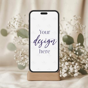 Op de afbeelding: Een zwarte smartphone mockup op een houten standaard met witte bloemen en groen op de achtergrond. Het telefoonscherm toont de tekst "Your design here".