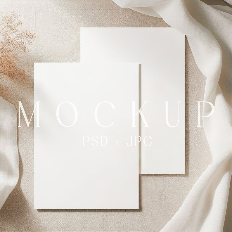 May include: Two blank white cards stacked on top of each other on a beige surface with a white fabric draped over the side. The text "MOCKUP PSD + JPG" is printed in white on the surface.