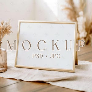 May include: A gold-framed picture frame with the word "MOCKUP" and "PSD + JPG" printed on the white surface. The frame is propped up on a wooden surface with a beige cloth and a vase with dried flowers.