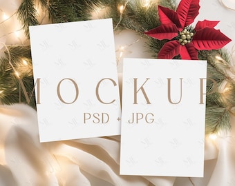 May include: Two white rectangular cards with the word "MOCKUP" and "PSD + JPG" printed on them. The cards are surrounded by Christmas greenery, fairy lights, and a red poinsettia flower. The overall theme is a holiday or seasonal mockup.