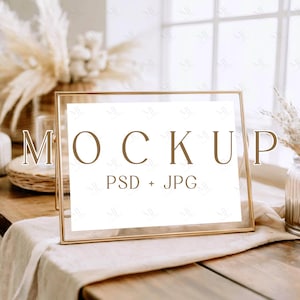 Op de afbeelding: Een goudkleurige rechthoekige lijst met het woord "MOCKUP" in bruine letters. De tekst "PSD + JPG" staat eronder. De lijst staat op een houten tafel met een beige doek, met gedroogde planten op de achtergrond.