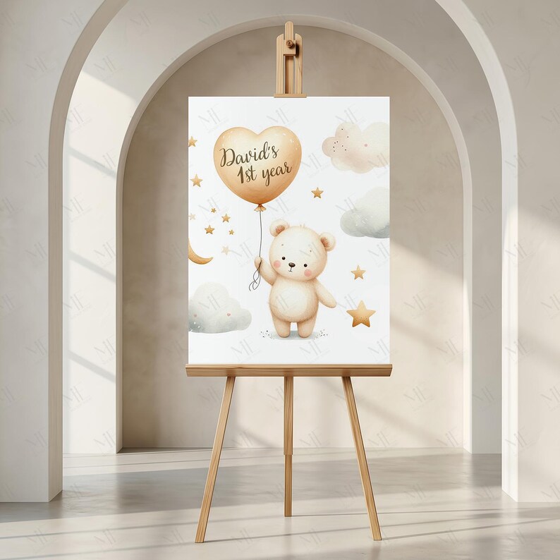 May include: A watercolour print on an easel depicting a teddy bear holding a heart-shaped balloon with the text "David's 1st year." The design includes stars, clouds, and a crescent moon in soft, neutral tones. The easel is made of light-coloured wood.
