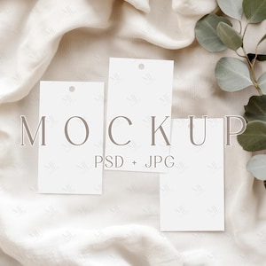 May include: Three blank white cards with rounded corners and a hole punched in the top corner, arranged on a white fabric background with a sprig of green eucalyptus leaves. The text "MOCKUP PSD + JPG" is printed in gray below the cards.