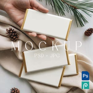 May include: A mockup image featuring several white rectangular objects with gold trim. One is held by a hand, while others are arranged on a beige cloth. Pinecones and a sprig of pine add to the aesthetic. The word "MOCKUP" is displayed.