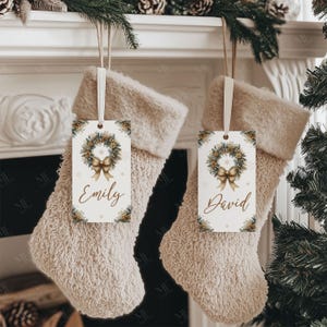 May include: Two beige Christmas stockings hang from a white mantelpiece, each with a decorative tag. The tags feature a wreath design with a gold bow and the names "Emily" and "David". The stockings are trimmed with a soft, fluffy material.