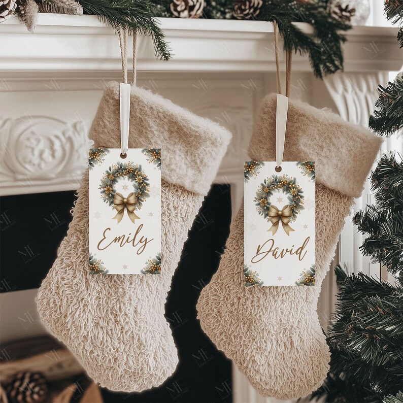 May include: Two beige Christmas stockings hang from a white mantelpiece, each with a decorative tag. The tags feature a wreath design with a gold bow and the names "Emily" and "David". The stockings are trimmed with a soft, fluffy material.