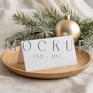 Może przedstawiać: Biała składana karta z napisem "MOCKUP" i "PSD + JPG", spoczywająca na okrągłym drewnianym talerzu. Złota ozdoba i gałązki sosny w tle, tworzące świąteczny motyw.