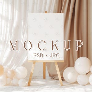 May include: A white canvas mockup on an easel with a wooden frame. The canvas is blank and ready for your design. The easel is surrounded by white and beige balloons and draped in white and beige fabric.