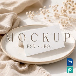 May include: A white rectangular folded card mockup resting on a round ceramic plate. The card features the text MOCKUP PSD + JPG in brown lettering. The plate is positioned near a beige fabric cloth and small white dried flowers.