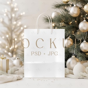 Könnte beinhalten: Eine weiße Einkaufstasche mit Griffen, mit dem Wort "MOCKUP" und "PSD + JPG" in einer neutralen Schriftart. Die Tasche steht vor einem Weihnachtsbaum mit goldenen und silbernen Kugeln und einer Geschenkbox.