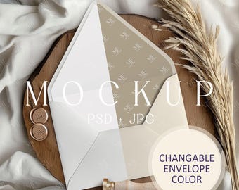 Op de afbeelding: Een witte envelop mockup met een beige gepatenteerde flap. De envelop ligt op een houten oppervlak met een gouden waszegel en een decoratieve veer. De tekst "MOCKUP PSD + JPG" en "CHANGEABLE ENVELOPE COLOR" is zichtbaar op de afbeelding.
