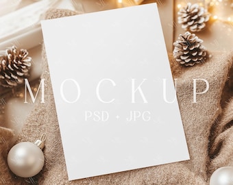 May include: A white card mockup on a brown textured surface with pine cones and string lights. The card is blank and has the text "MOCKUP PSD + JPG" printed on it.
