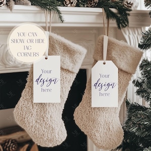 May include: Two beige Christmas stockings with white tags that read "Your design here." The stockings are hanging from a white mantelpiece decorated with pine needles and pine cones. The text "YOU CAN SHOW OR HIDE TAG CORNERS" is in a circle.
