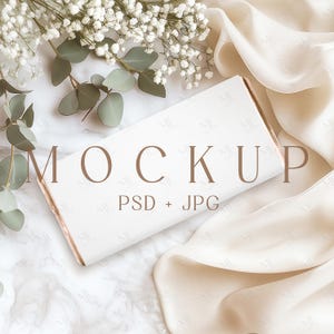 Könnte beinhalten: Ein weißer Mockup einer Produktverpackung mit dem Text "MOCKUP PSD + JPG" auf einer weißen Marmorfläche mit einem weißen Seidenstoff und einem Zweig weißer Blumen.