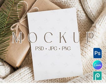 May include: A white mockup card with the word "MOCKUP" in a serif font, placed on a cream-coloured knitted blanket. A wrapped gift with twine and a sprig of evergreen is next to the card. The text "PSD + JPG + PNG" is below the word "MOCKUP".