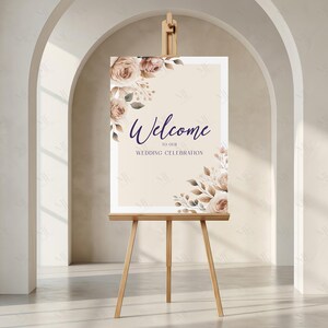 May include: A wooden easel displays a wedding welcome sign. The sign features the word "Welcome" in elegant script, with "to our WEDDING CELEBRATION" below. Floral arrangements in shades of beige and brown adorn the corners of the sign.