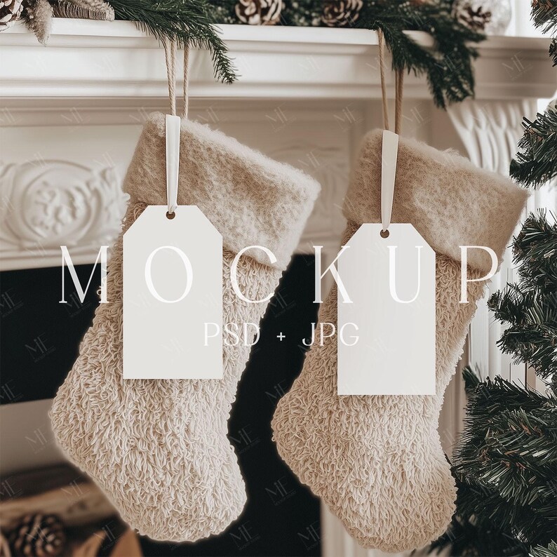 May include: Two beige Christmas stockings with blank white tags hanging from a white mantelpiece. The stockings have a soft, textured appearance. The tags are suspended by thin, light-colored ribbons. The background includes pine branches and pine cones.