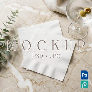 Puede incluir: Dos servilletas de papel blancas con un borde texturizado se muestran sobre una superficie de mármol. La palabra "MOCKUP" está impresa en las servilletas. También se ve una copa de martini con aceitunas y una ramita de eucalipto.