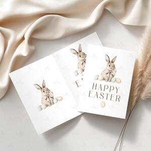 May include: Three white greeting cards with watercolor illustrations of Easter bunnies wearing pink bows and surrounded by pastel eggs. One card reads "HAPPY EASTER" in brown text. The cards are arranged on a cream-colored surface.