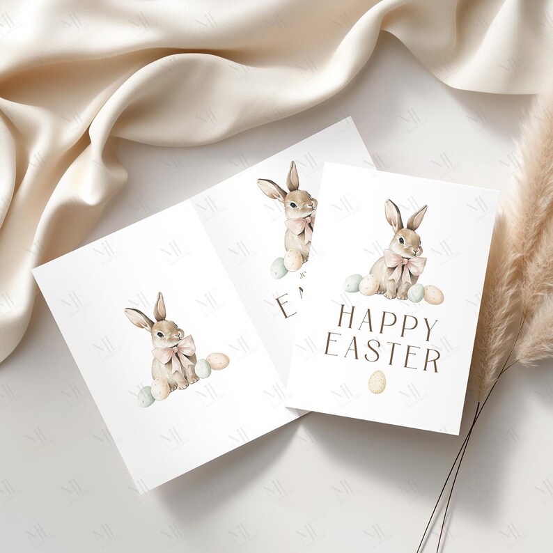 May include: Three white greeting cards with watercolor illustrations of Easter bunnies wearing pink bows and surrounded by pastel eggs. One card reads "HAPPY EASTER" in brown text. The cards are arranged on a cream-colored surface.