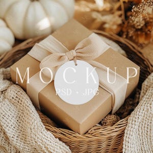 May include: A brown gift box tied with a cream-colored ribbon and bow, with a white circular tag. The box is in a woven basket with a cream-colored blanket. The word "MOCKUP" is printed on the tag, with "PSD + JPG" below.