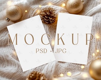 May include: Two white rectangular cards with the word "MOCKUP" in brown letters. The cards are surrounded by gold Christmas ornaments, pine cones, and a soft, white knitted blanket. The text "PSD + JPG" is below the word "MOCKUP".