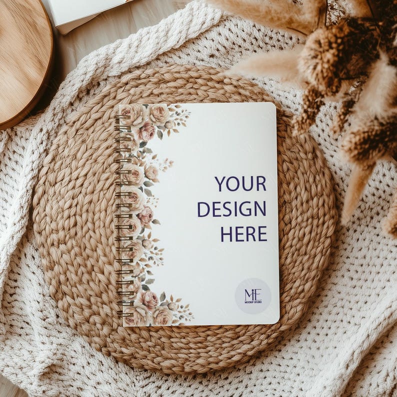 May include: A white notebook with a floral design on the left side and the text "YOUR DESIGN HERE" in the center. The notebook is on a woven straw mat and a white knitted blanket.