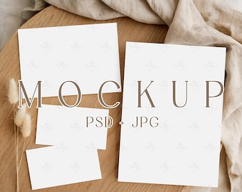 May include: A mockup scene featuring blank white cards of various sizes arranged on a wooden surface. The word "MOCKUP" is displayed in a brown font, with "PSD + JPG" below. Dried flowers and a beige fabric are also present.