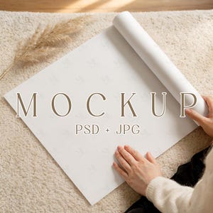 Puede incluir: Una maqueta de papel blanco con la palabra "MOCKUP" en letras marrones. El papel está parcialmente enrollado y se ven las manos de una persona sujetando el rollo. El fondo es una alfombra beige y una planta seca está en la esquina superior izquierda.