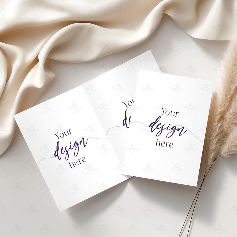 May include: Three white greeting cards with the text "Your design here" in a handwritten font. The cards are arranged on a white surface with a beige fabric and dried ornamental grass in the background.