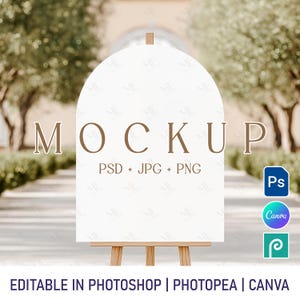 Puede incluir: Una maqueta de cartel blanco en forma de arco sobre un caballete con el texto "MOCKUP PSD + JPG + PNG" en texto marrón. El cartel está delante de un fondo borroso de árboles y un edificio.