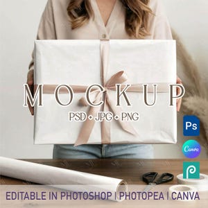 Op de afbeelding: Een witte geschenkdoos verpakt met een satijnen lint en strik. Het woord "MOCKUP" staat op de voorkant. De afbeelding bevat ook een rol wit papier, een schaar, plakband en pictogrammen voor Photoshop, Canva en Photopea.