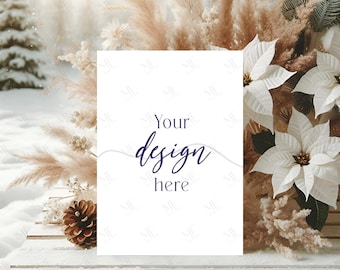 May include: A white card with the text "Your design here" in a dark blue script font. The card is propped up on a white wooden surface, with a backdrop of winter foliage, including white poinsettias and pampas grass.