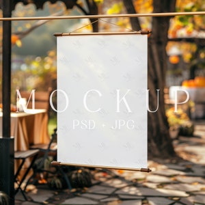 May include: A white canvas banner mockup hanging from a gold rod in a fall setting. The banner is blank and ready for your design. The background is blurred and features a rustic outdoor scene with fall foliage and pumpkins.