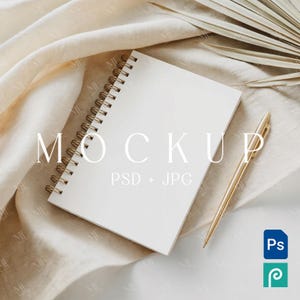 Op de afbeelding: Een wit spiraalgebonden notitieboek met goudkleurig draad en een gouden pen ligt op een crèmekleurige stof. Het woord "MOCKUP" staat op het notitieboek. De tekst "PSD + JPG" staat eronder.