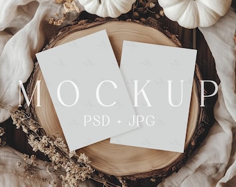 May include: A mockup scene featuring two light gray cards with the word "MOCKUP" in white. The cards rest on a wooden slice, surrounded by white pumpkins, dried flowers, and a beige fabric.