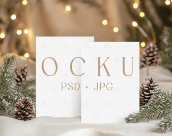 May include: Two white cards with the text "MOCKUP PSD + JPG" on a white fabric background with pine branches and pine cones. The background is blurred with warm white lights.