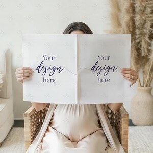 Puede incluir: Una persona sentada en una silla de mimbre sostiene dos papeles cuadrados blancos. Cada papel tiene el texto "Your design here" en una fuente de escritura morada oscura. El fondo incluye un vestido beige, un jarr&oacute;n y plantas secas.