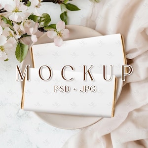 May include: A white mockup of a chocolate bar with gold foil edges, on a white plate with a floral arrangement. The text "MOCKUP PSD + JPG" is printed on the chocolate bar.