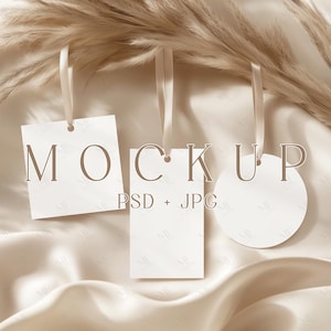 Puede incluir: Tres etiquetas blancas de maqueta con cintas color crema cuelgan sobre un fondo de tela beige y hierba ornamental seca. Las etiquetas son cuadradas, rectangulares y circulares, con la palabra "MOCKUP" y "PSD + JPG" impresas.