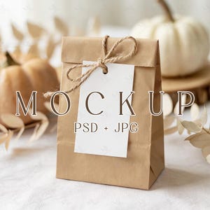 Pode incluir: Uma sacola de papel castanho com uma etiqueta branca amarrada com barbante. A etiqueta diz "MOCKUP PSD + JPG". O fundo inclui abóboras e folhagens secas, sugerindo um tema de outono.
