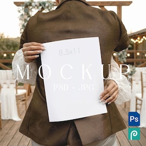 May include: A white 8.5x11 inch mockup held by a person wearing a brown blazer and white blouse. The word "MOCKUP" is printed on the paper. The background includes a wooden structure and chairs, suggesting an outdoor event.