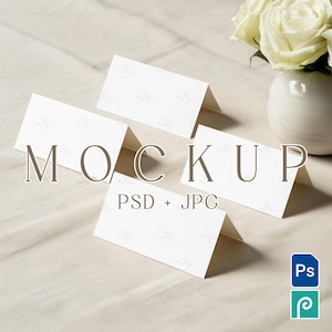 Puede incluir: Cuatro tarjetas blancas plegadas en forma de tienda de campaña sobre un mantel beige. La palabra "MOCKUP" está impresa en marrón sobre las tarjetas. El texto "PSD + JPG" está debajo. Un pequeño jarrón blanco con rosas blancas en el fondo.